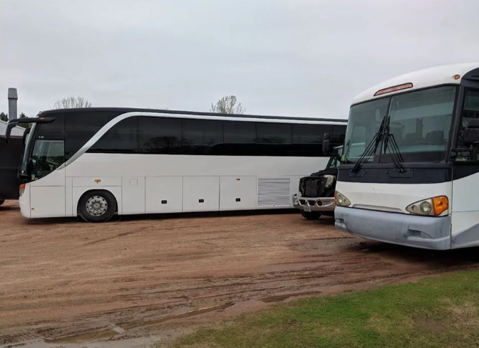 White coach bus parked on a dirt lot beside a gray bus and other vehicles
