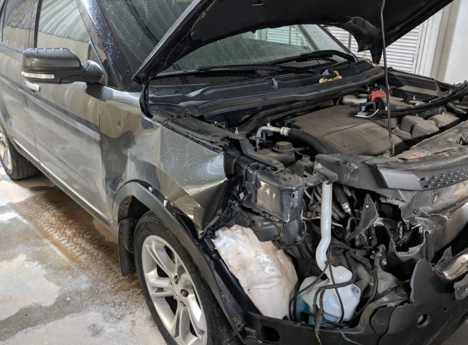 Front-left view of a silver car with severe collision damage and a crumpled hood in a garage.