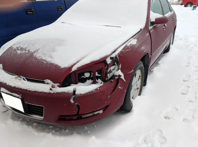 Maroon car with front-end damage, parked in snow with snow covering the hood.