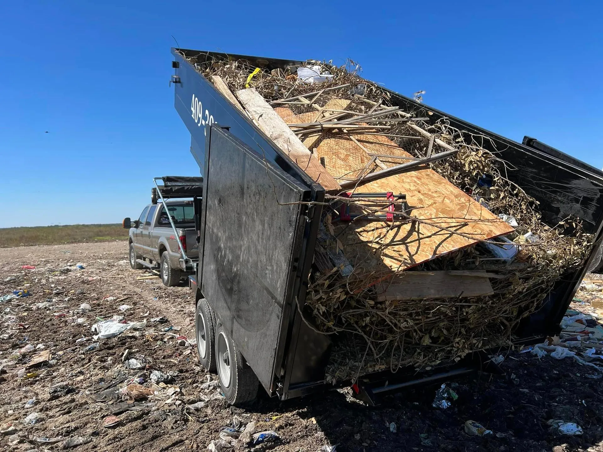 A dumpster filled with wood is sitting in a field next to a truck.