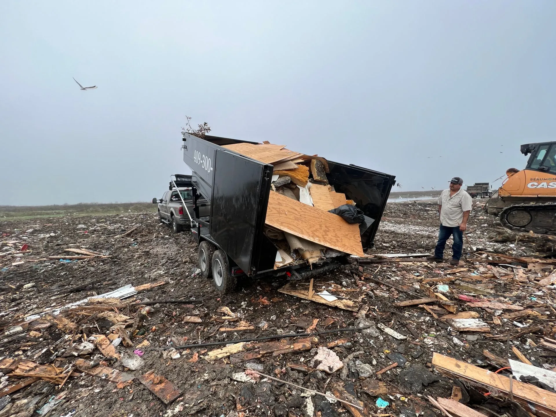 A man is standing next to a trailer filled with junk in a pile of trash.