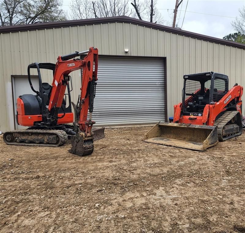 Two orange Kubota construction vehicles parked outside a beige metal building.