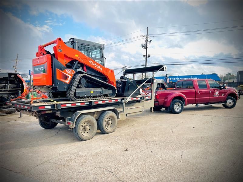 Red truck towing a trailer with an orange Kubota track skid steer.