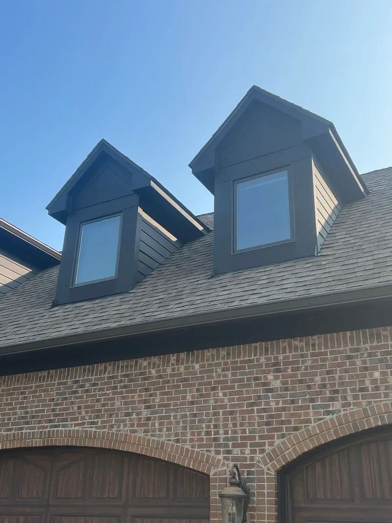 Two dark dormers with windows on a brick building with a brown shingled roof, against a blue sky.