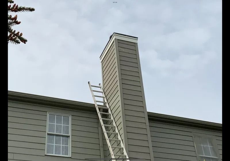 Ladder leaning against a tall chimney on a gray house under a cloudy sky.