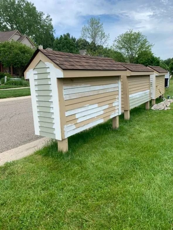 Row of beige and white mailbox structures with brown shingle roofs, standing on posts beside a green lawn and road.