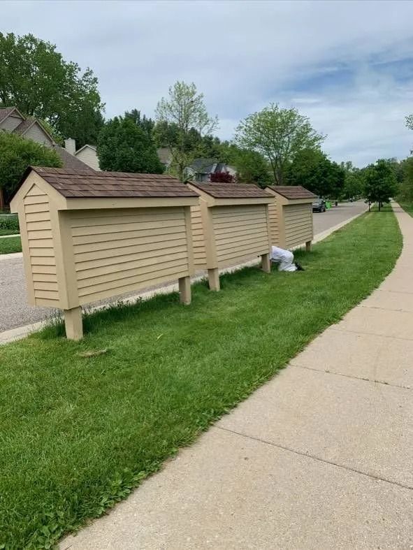 Three wooden structures with brown roofs on a grassy verge next to a sidewalk.