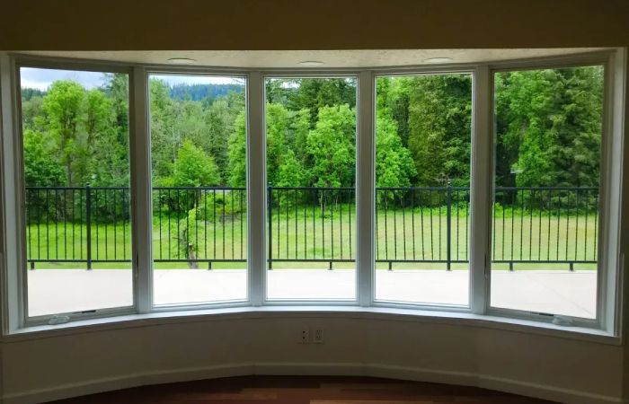 Curved bay window with white frame overlooking a green wooded area and black iron railing.