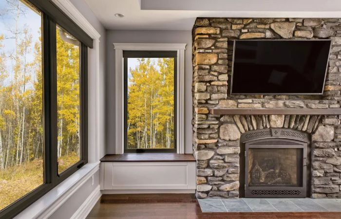 Living room with stone fireplace, TV, and windows overlooking fall trees.
