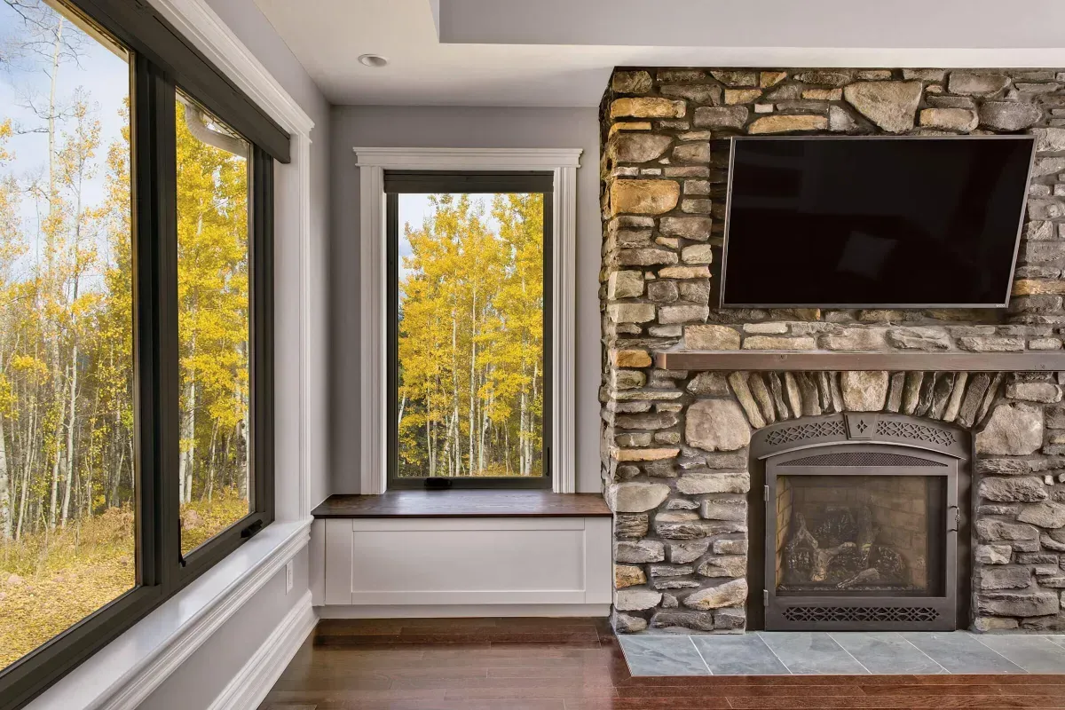 Living room with stone fireplace, TV, and windows overlooking fall trees.