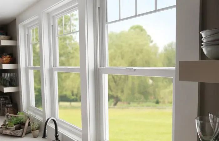 White double-hung windows overlook a green lawn and trees. Kitchen shelving with dishes is visible.