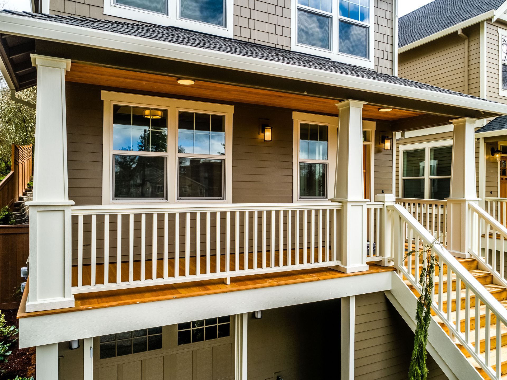 Two-story house with a covered porch, white columns, and wooden railings. Brown siding with a garage below.