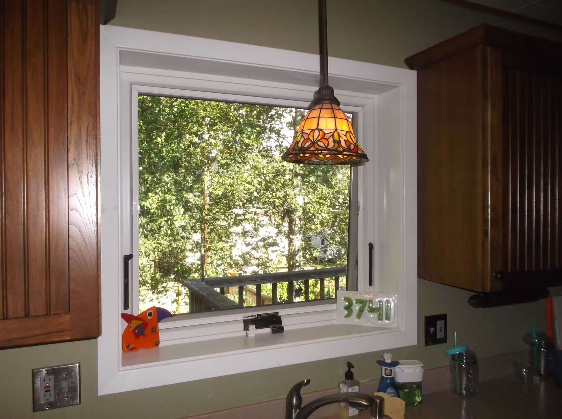 White-framed kitchen window with view of trees and deck, flanked by brown cabinets and a hanging lamp.
