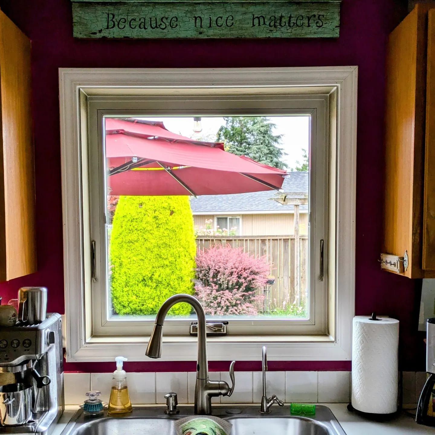 Kitchen sink and window overlooking a yard with red umbrella and green and pink bushes.