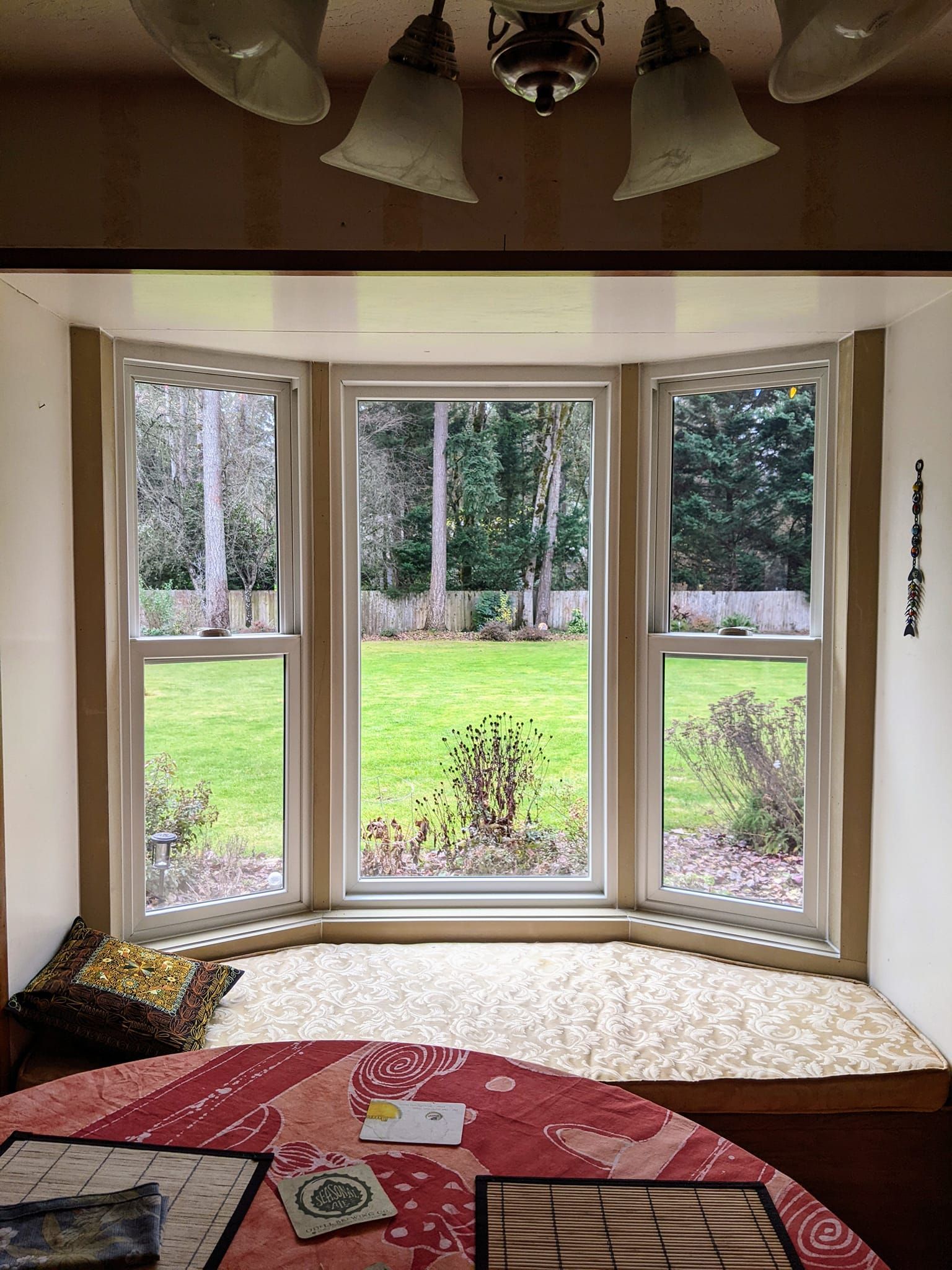 Bay window overlooking a green lawn and trees. A seating area with cushion and a table in the foreground.