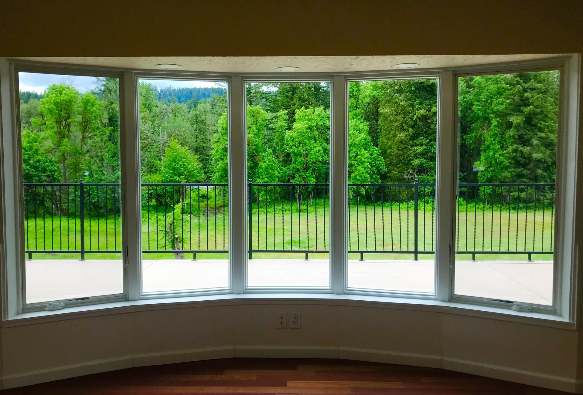 Bay window overlooking a lush green landscape with a black railing, set in a light-filled room with wood flooring.