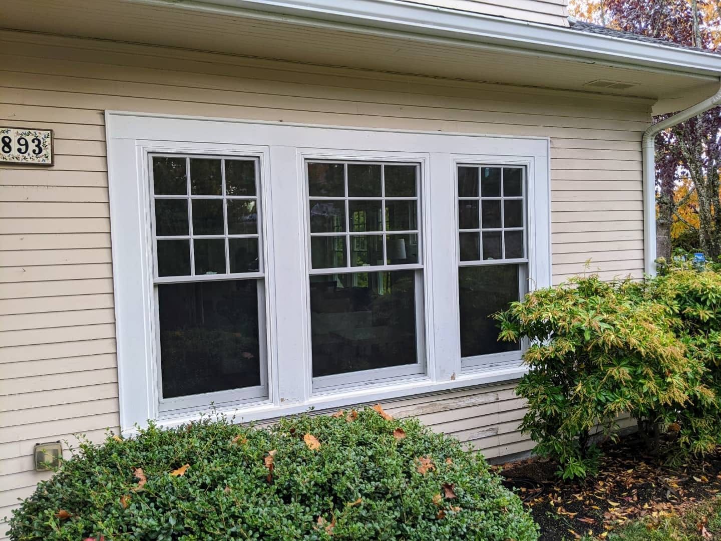 Three white-framed windows on a beige house exterior with bushes in front. A house number is visible.