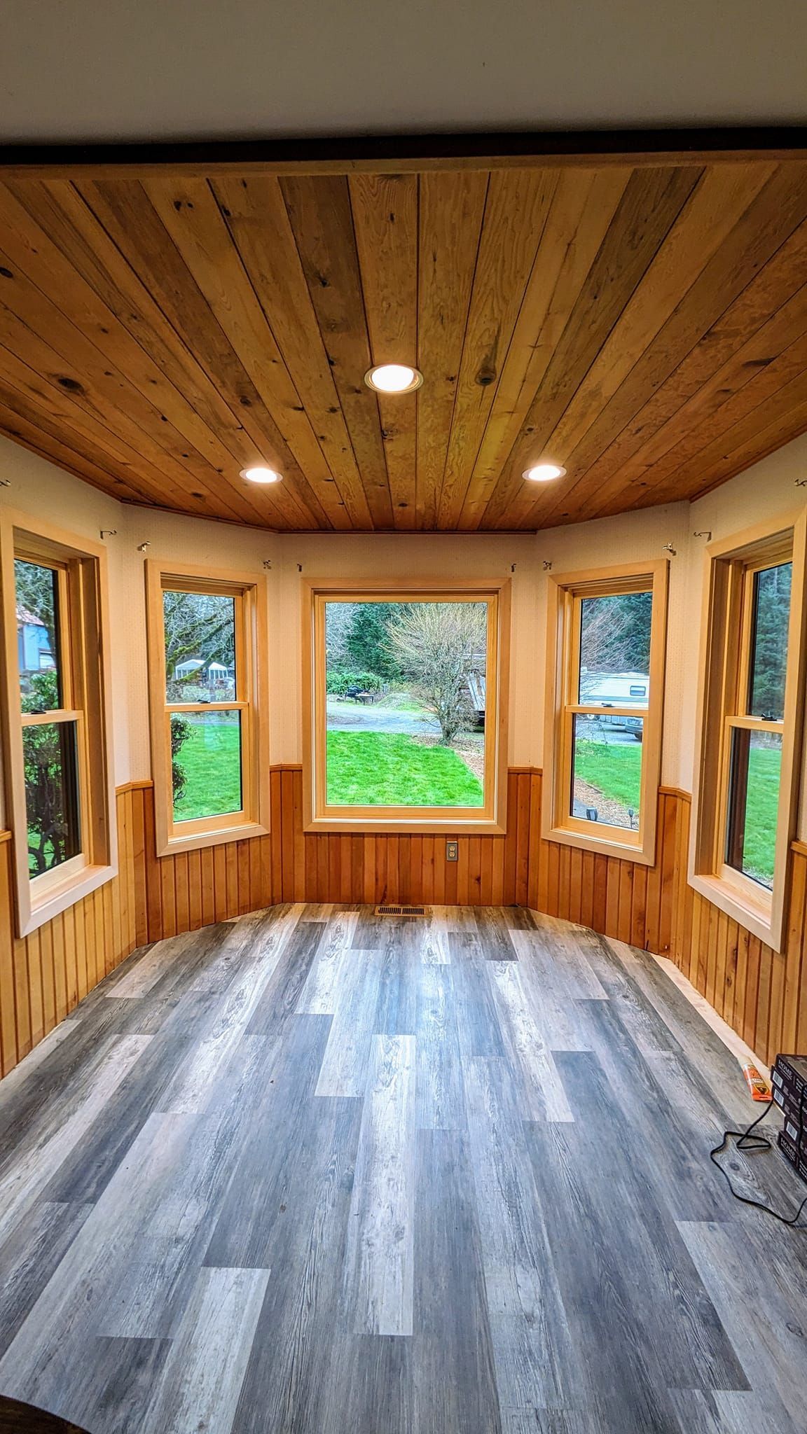 Bay window room with wood paneling on walls and ceiling, gray wood-look floor, and three windows overlooking a green yard.
