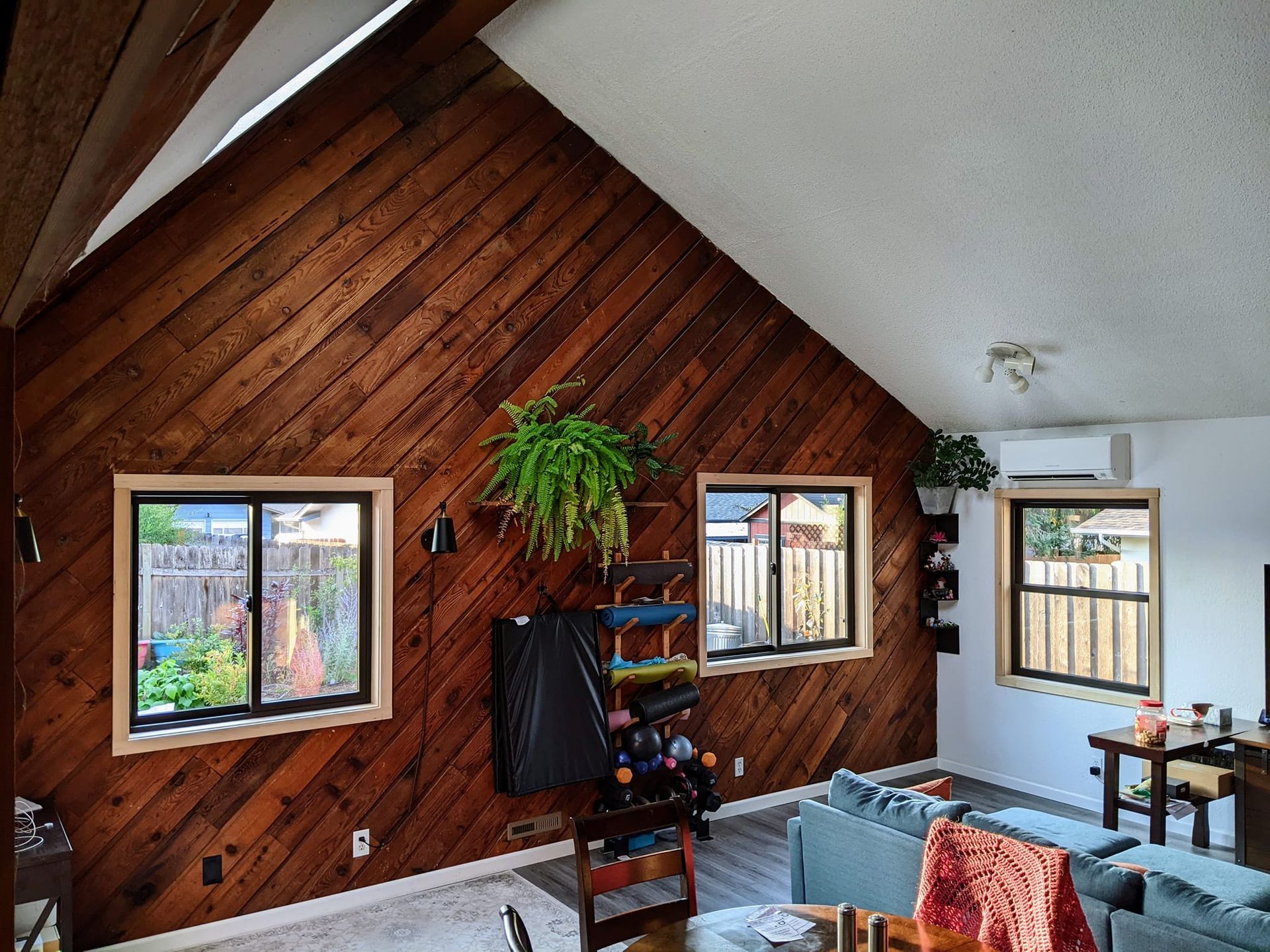 Interior room with wood-paneled wall, windows, and a hanging fern. A couch and other furniture are visible.