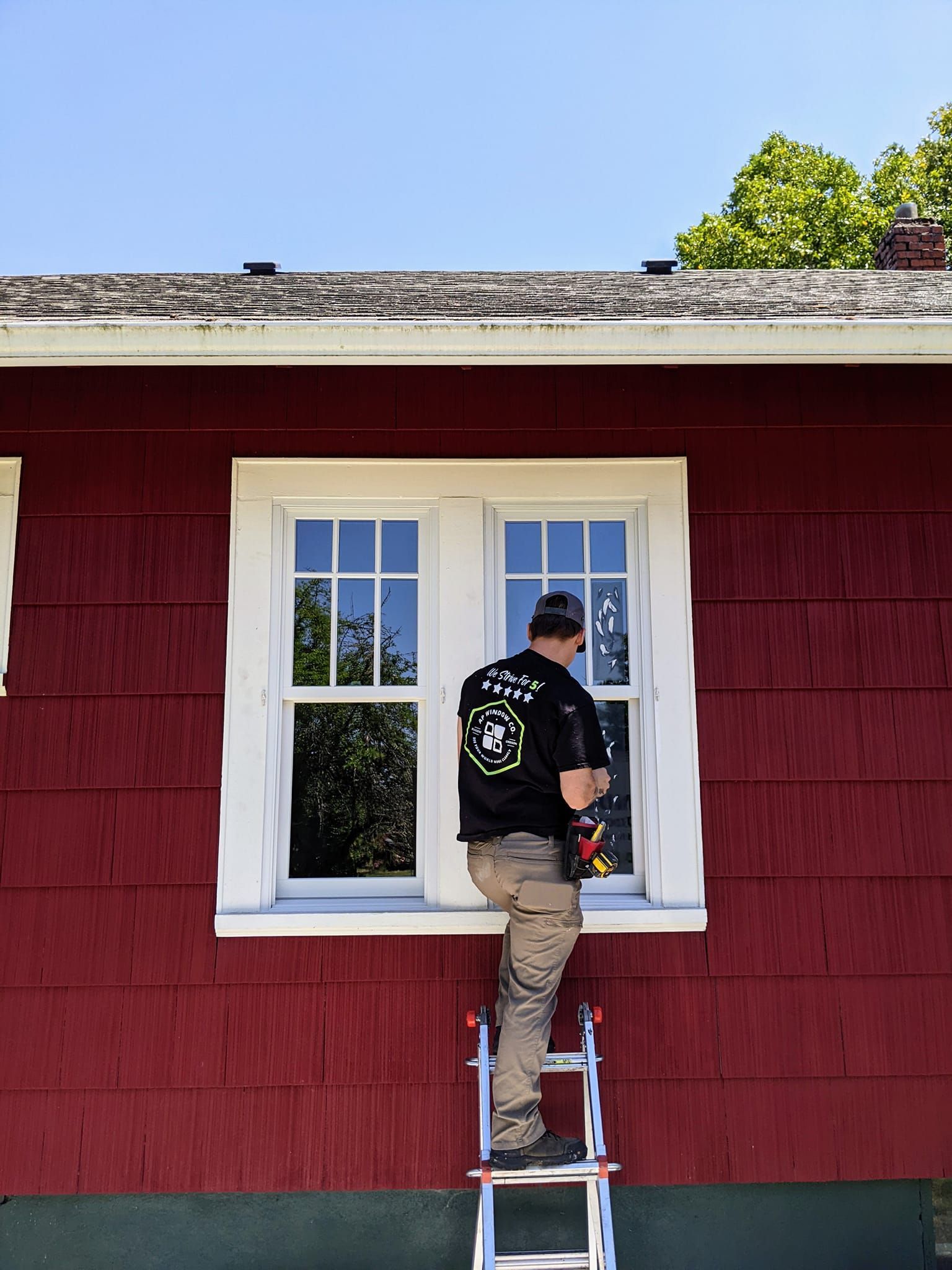 Person on a ladder cleaning a window of a red house with white trim against a blue sky.