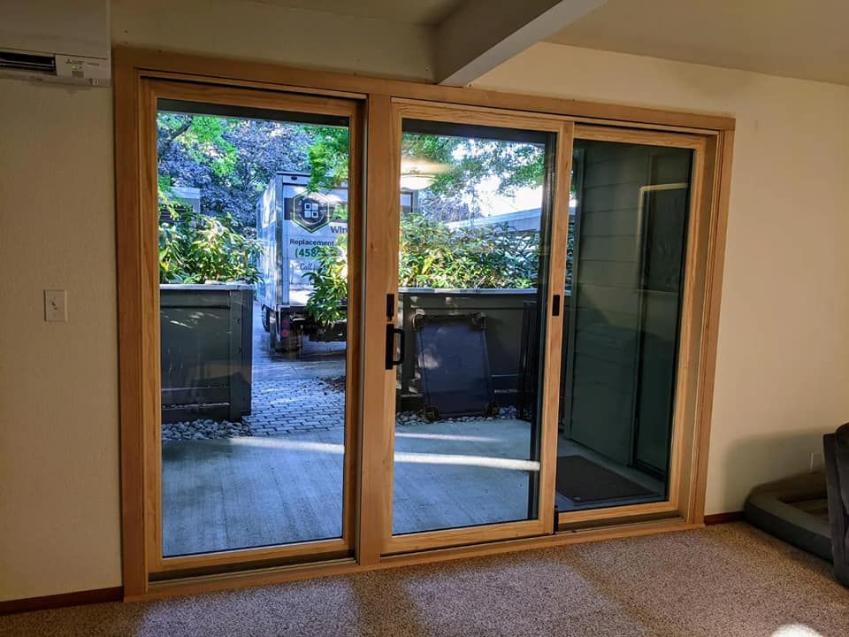 Sliding glass doors with wooden frames open to an outdoor patio with greenery.