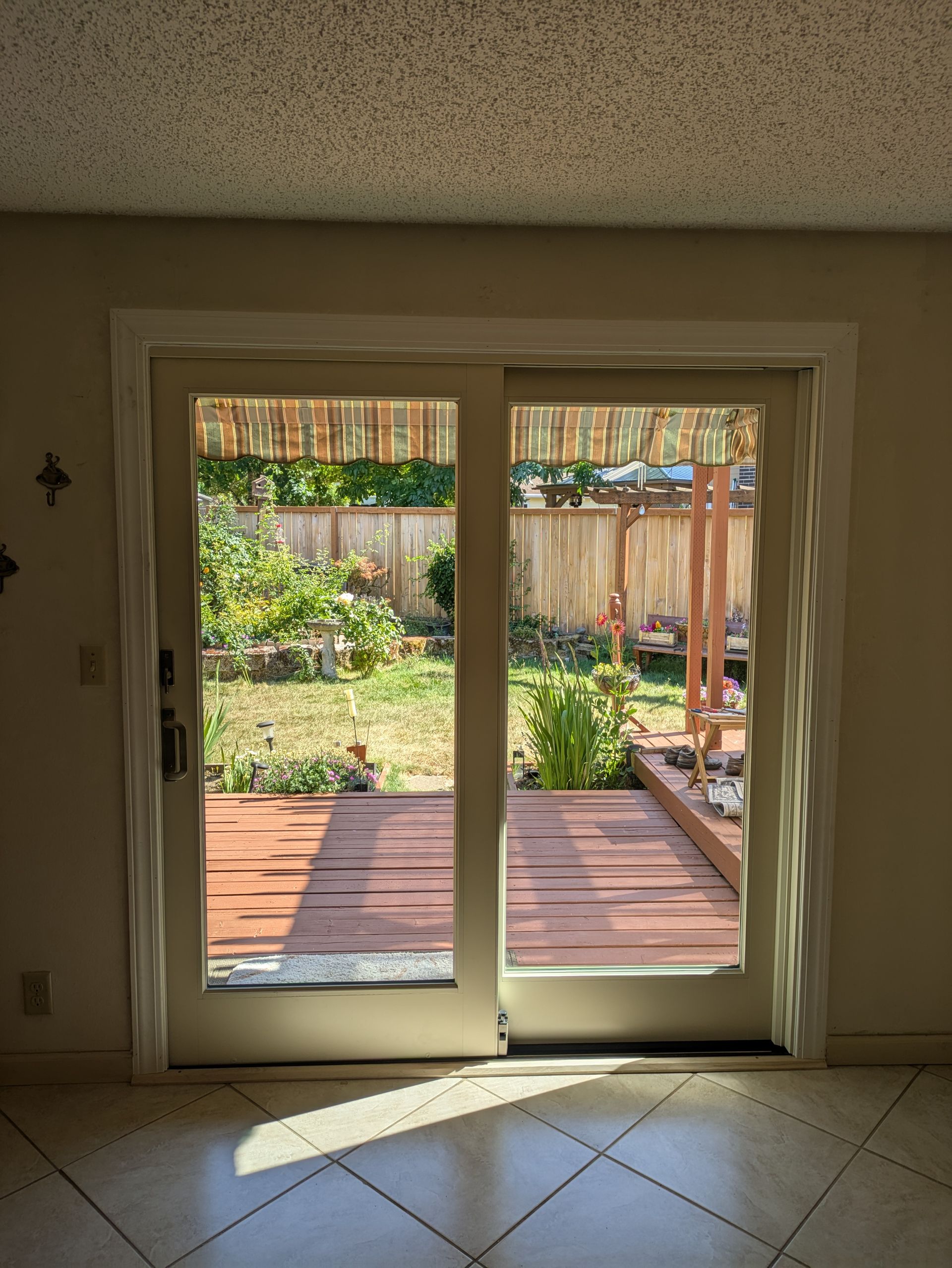 Sliding glass doors open onto a sunlit backyard with a wooden deck and fence. The interior has a tiled floor.