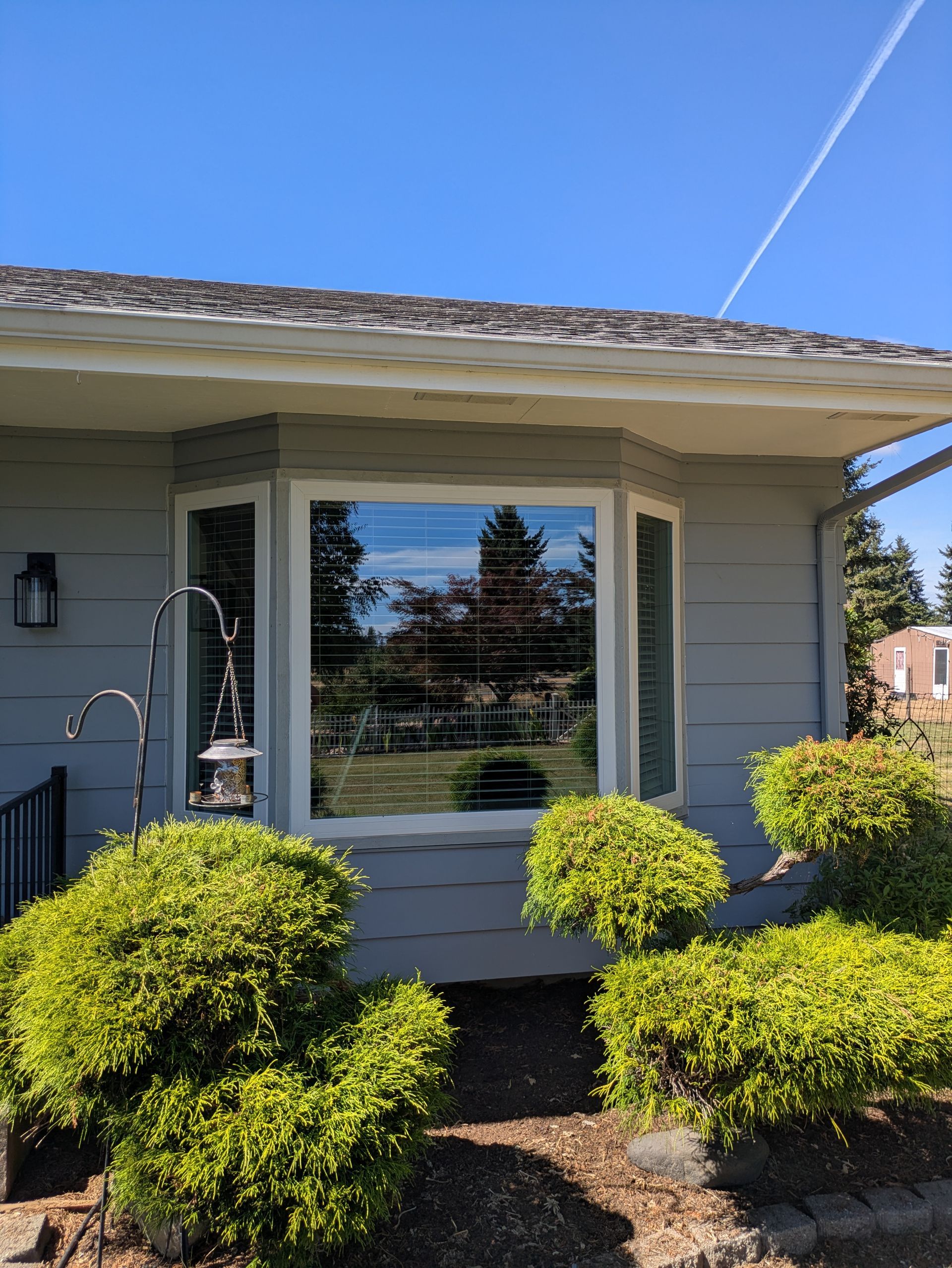 Bay window on a light gray house with green bushes and blue sky reflection.