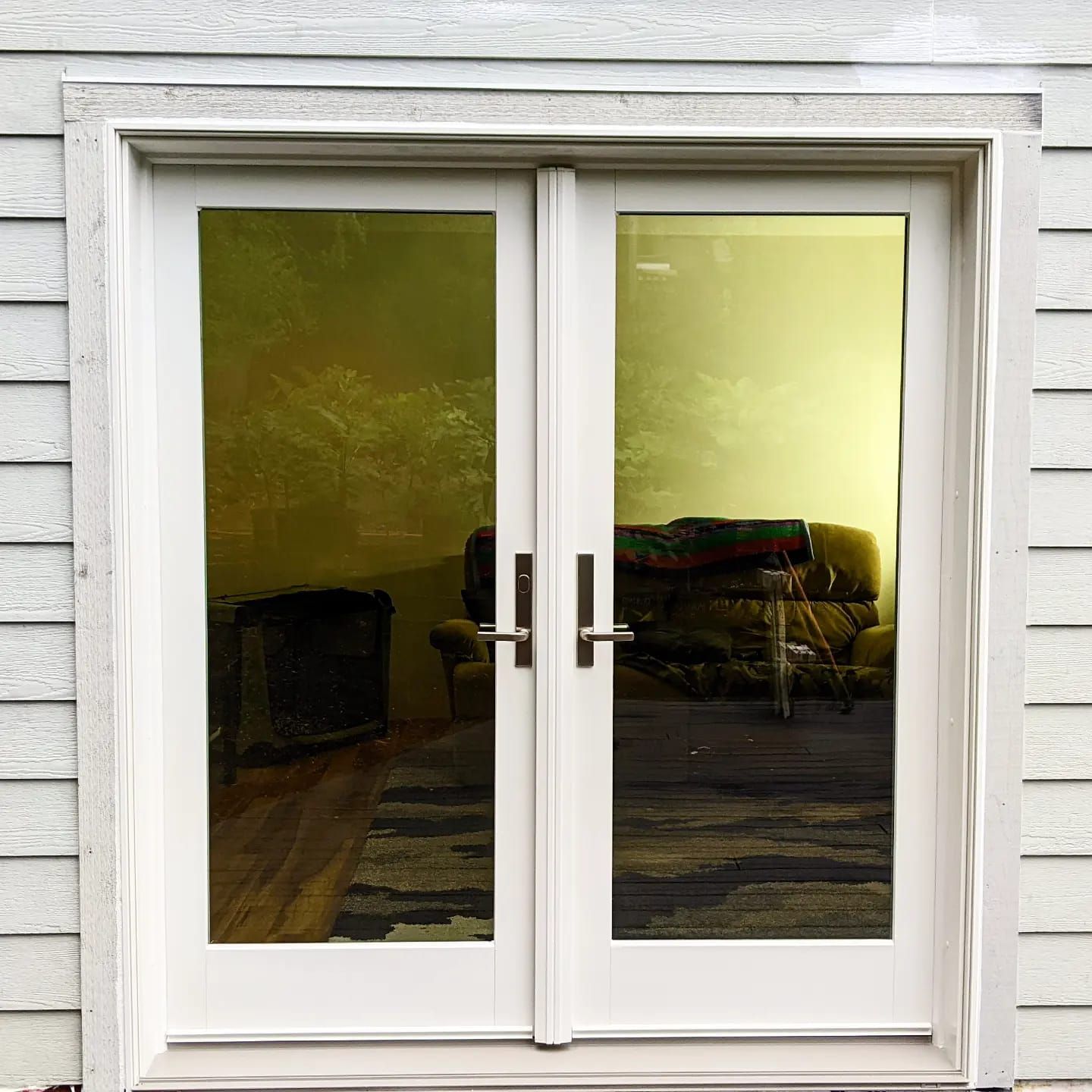 White French doors reflect a room with furniture and sunlight. Exterior has white siding.