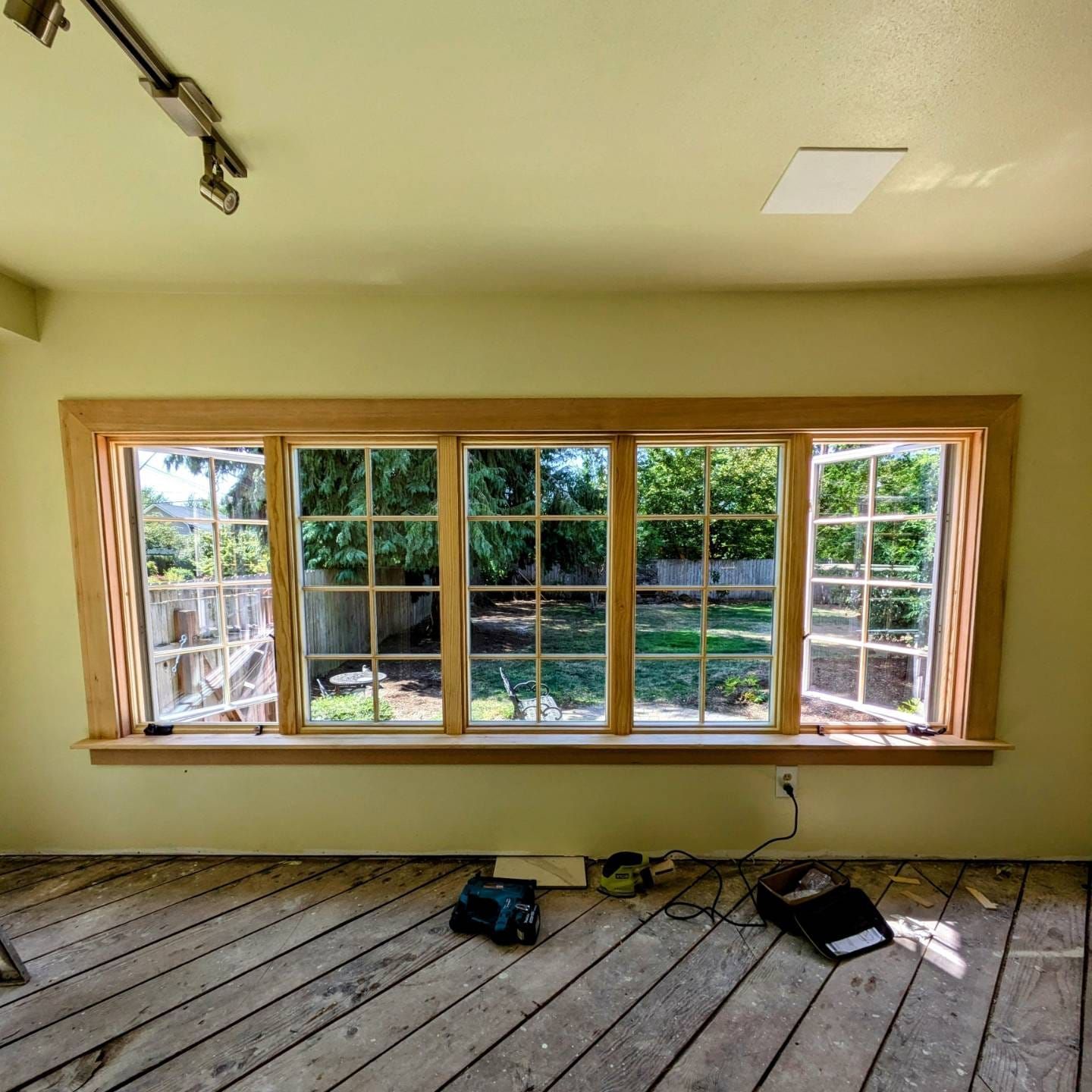 Room interior with light wood-framed windows open to a backyard. Unfinished floor, tools.