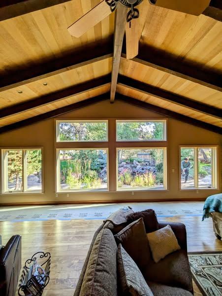 Living room with wooden ceiling, large windows overlooking greenery, and a brown sofa.