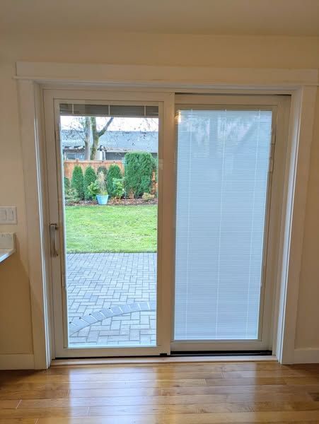 Sliding glass door with white frame and blinds. View of a backyard with green grass, brick patio.