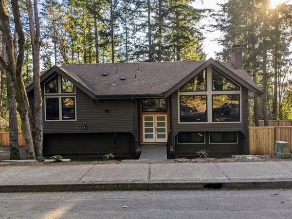 Brown house with large windows, set against a backdrop of tall trees and a sidewalk.