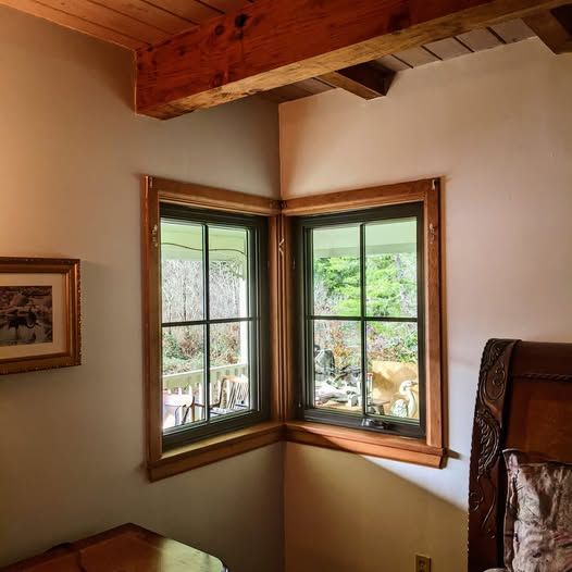 Corner window with wooden frame, overlooking a green outdoor scene. Interior features a beamed ceiling and a bed.