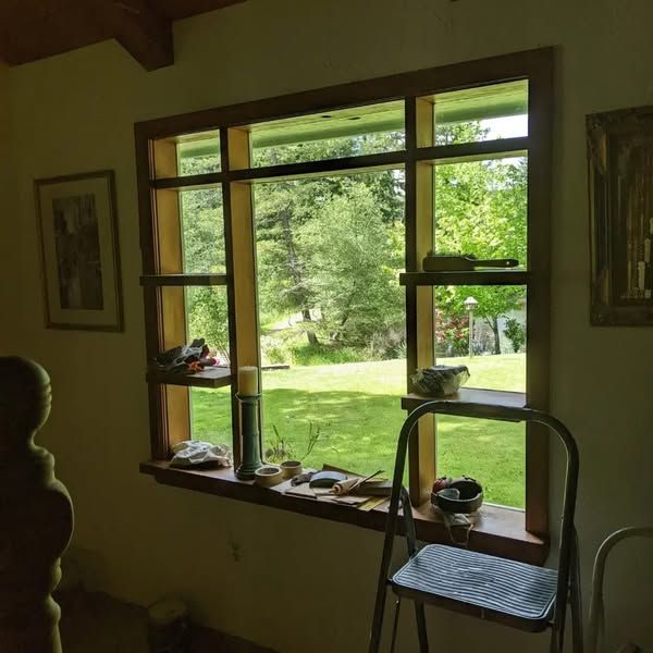 Wooden window overlooking a green yard, tools and supplies on the sill. A ladder sits below.