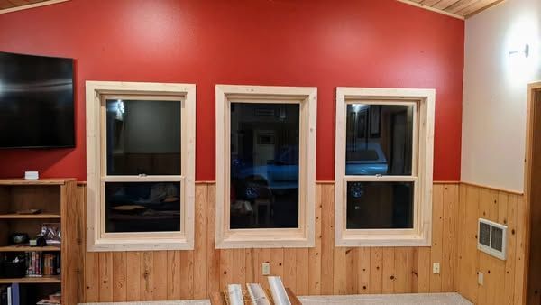 Three windows with light wood trim on a red wall above wood paneling.