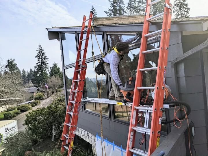 Man on platform outside house using saw on window frame, orange ladders, safety harness, and scenic background.