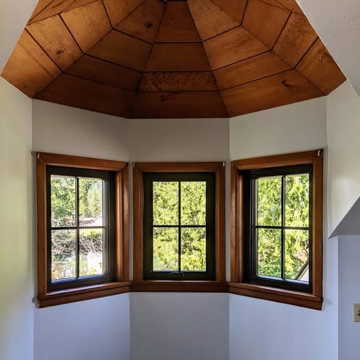 Three windows in a corner with a wooden ceiling. Lush green trees are seen through the windows.