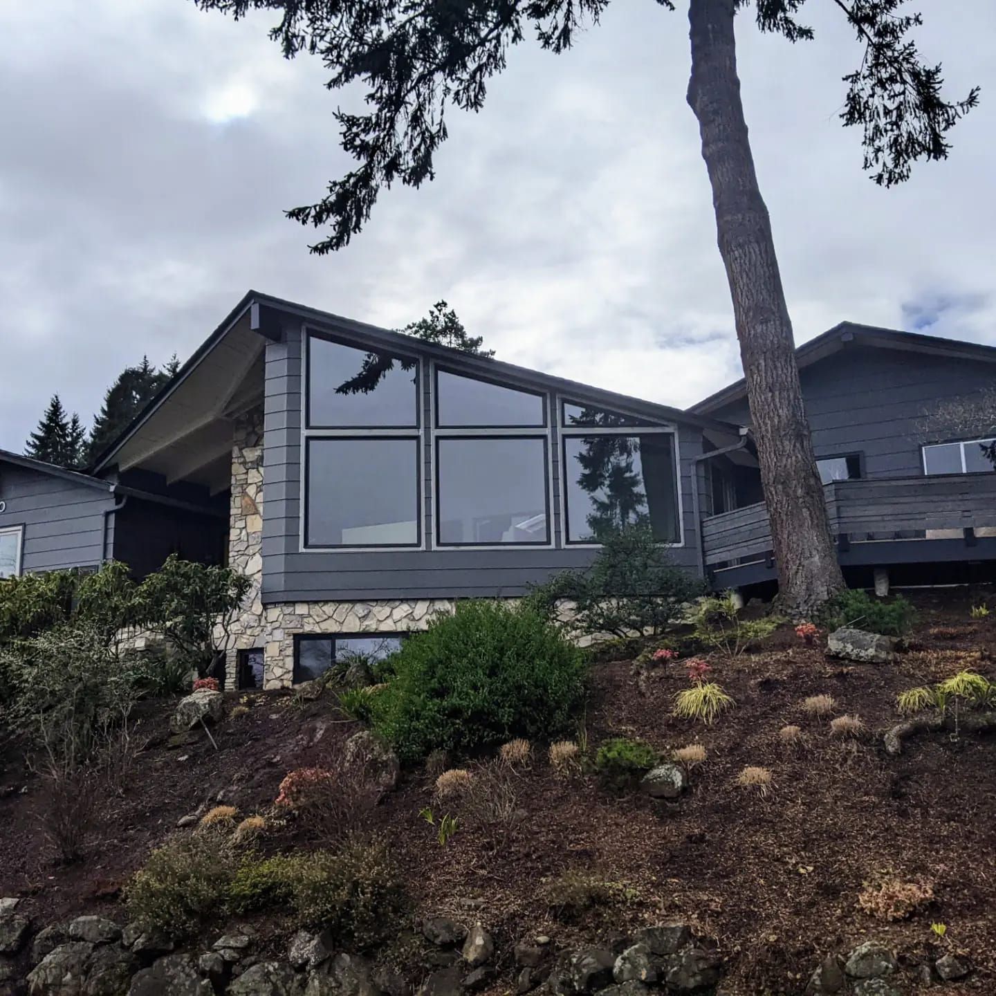 Modern gray house with angled roof, large windows, and rock and wood exterior, on a hillside.