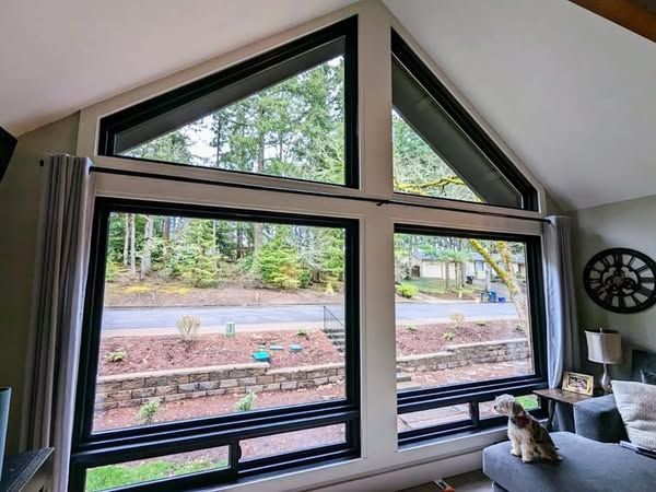 Large windows with black frames. Two rectangular lower windows and two triangular upper windows offer a view of trees and a road.