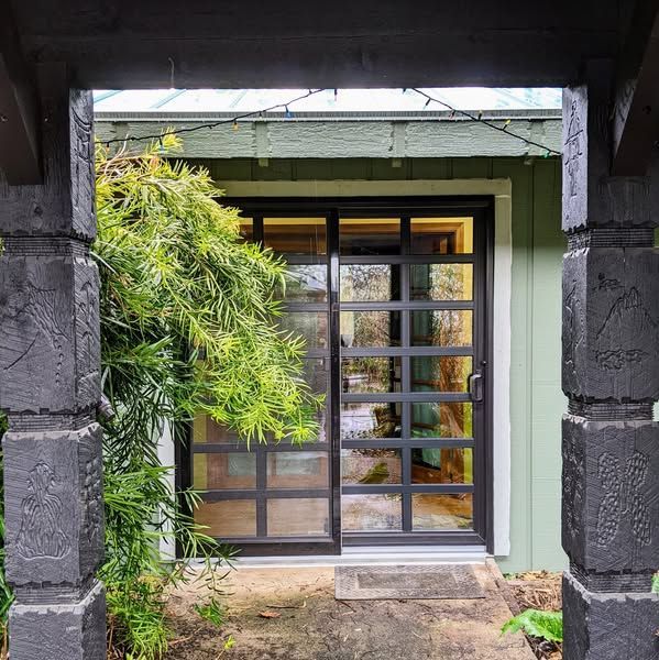 Black-framed glass sliding door of a green building viewed from a dark wooden structure; bamboo on the left.
