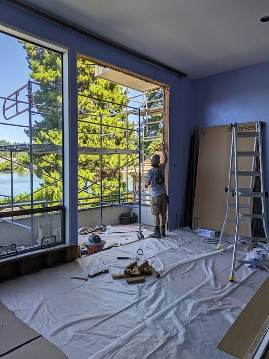 Man installing a window frame in a room with a scenic view, using scaffolding and tools; room has blue walls.