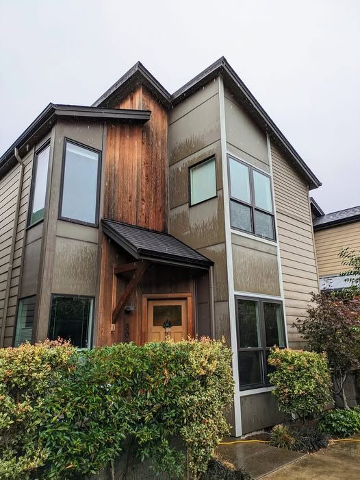 Two-story modern house with brown and gray siding, windows, and a wooden entrance. Green bushes in front.