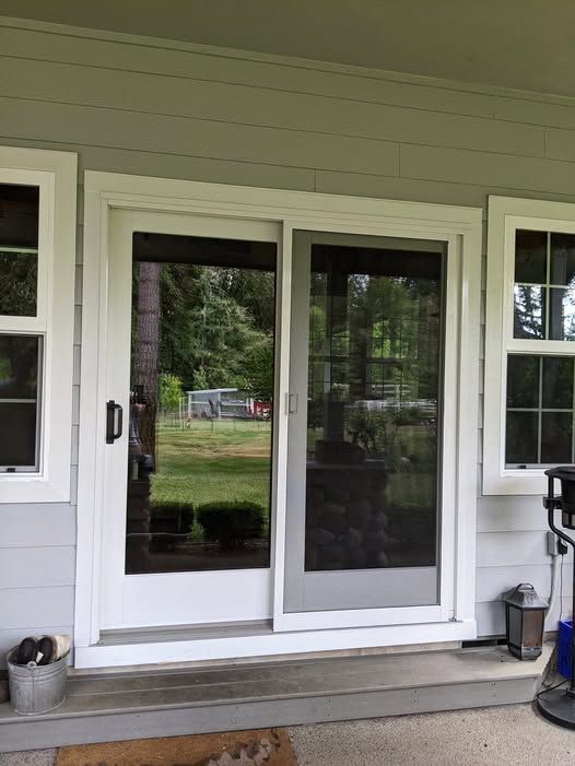 Sliding glass door with screen on a light gray house. White trim. Green yard reflected in glass.