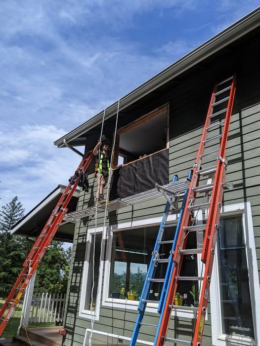 Person on ladders installing window on a green house.  Two red and one blue ladder support the work. Sunny day.