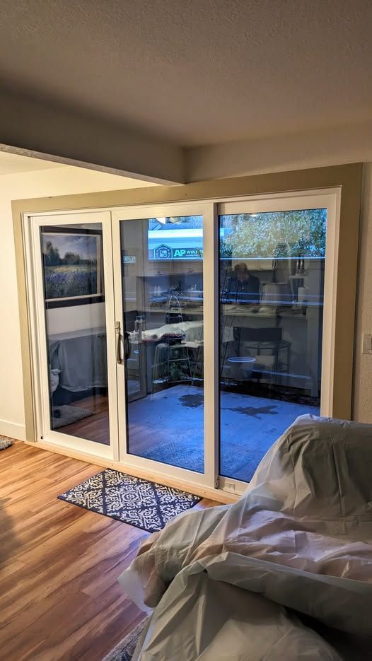 Sliding glass doors lead to a patio. Indoor view: wooden floor, couch. Outdoor view: gray patio, plants.