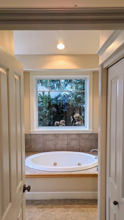 Bathroom with a jacuzzi tub under a window. Light beige walls, natural view outside.