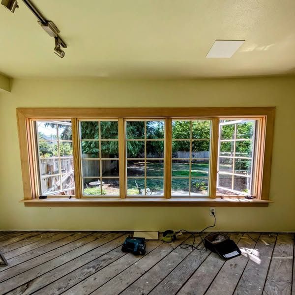 Room with a large window; wooden trim and frame. Old wood floor. Interior with a green backyard view.