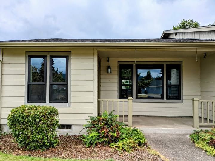 Beige house with dark-framed windows, porch, and greenery under a cloudy sky.