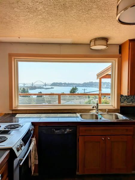 Kitchen with a large window overlooking a bridge and water; dark countertops, black appliances, and wooden cabinets.