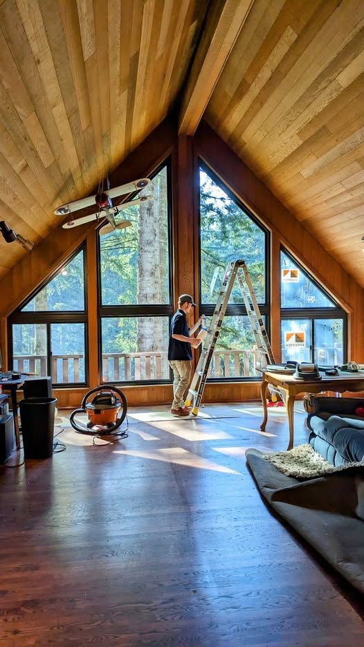 Person on ladder near large windows in wooden-paneled room. Vacuum cleaner and table visible.